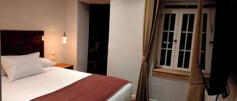 A hotel room with a large bed, a bedside table with a lamp, a window with curtains, and a wooden floor.