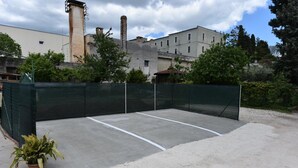 Una cancha de tenis exterior con una valla verde, una planta en maceta y edificios al fondo.