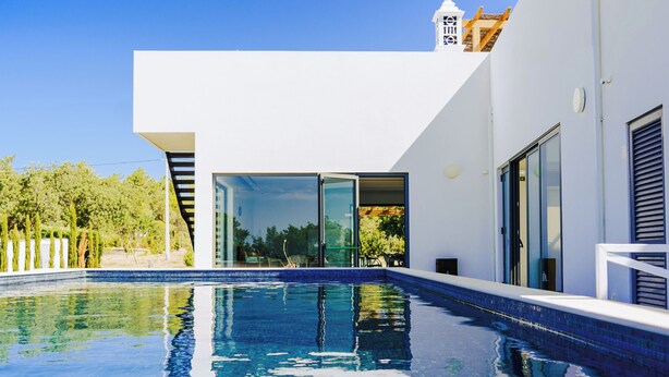 Un espace piscine moderne avec une piscine à l’eau d’un bleu limpide, un bâtiment blanc doté de grandes portes vitrées et une vue sur la verdure en arrière-plan.