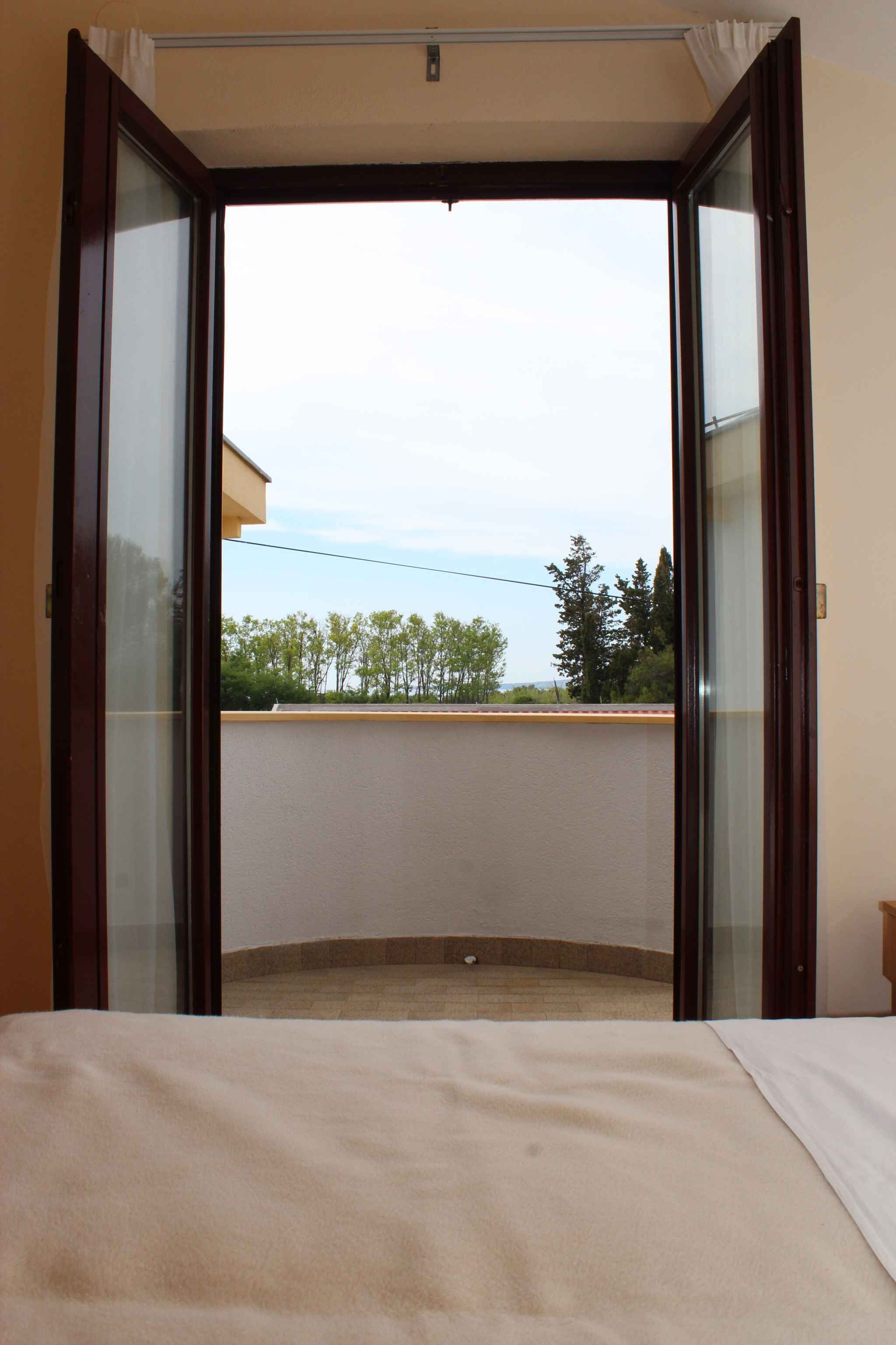A room with a bed, a sliding glass door leading to a balcony, and a view of trees and a building.
