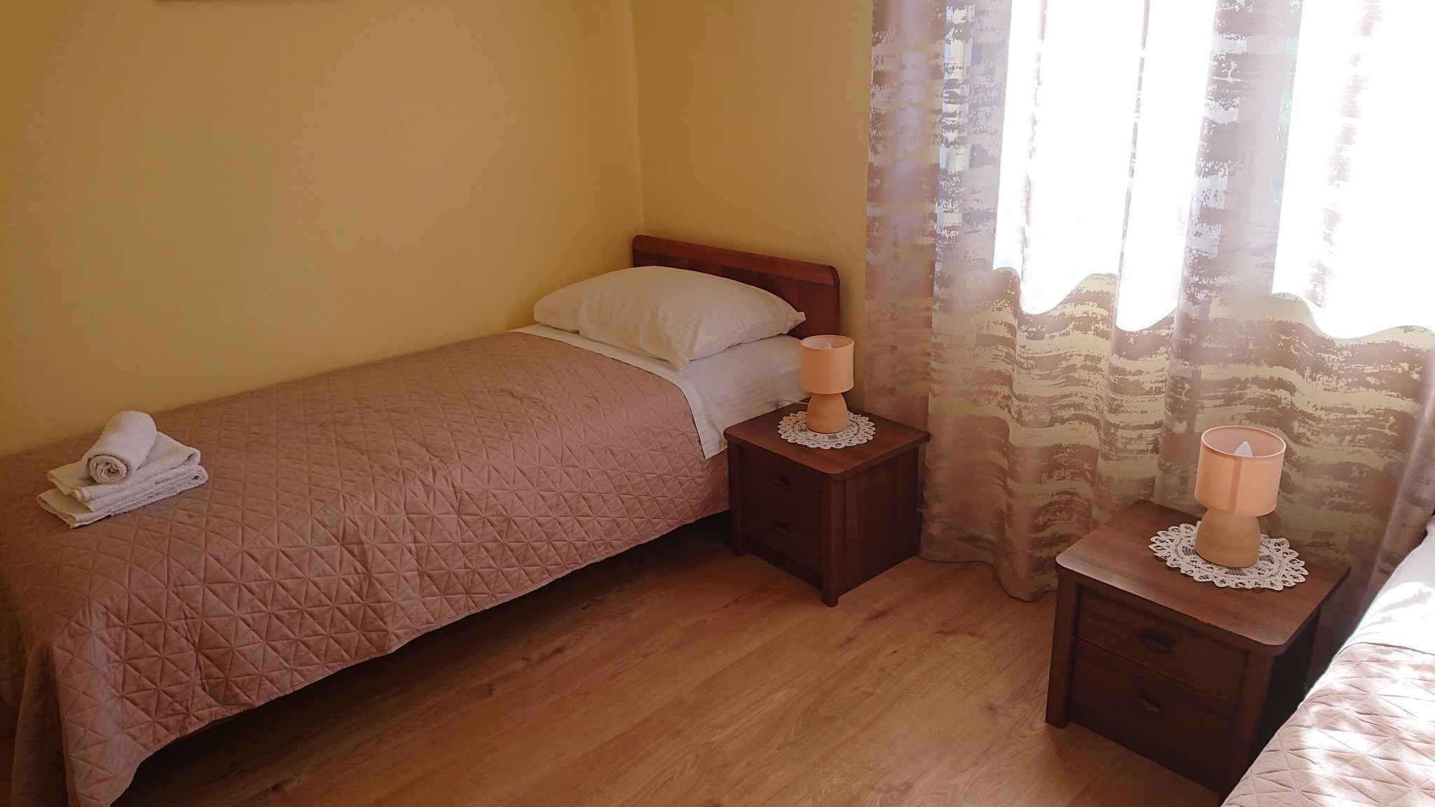 Un petit lit bien fait, recouvert d’une couverture marron, une table de chevet avec une lampe et une fenêtre avec des rideaux.