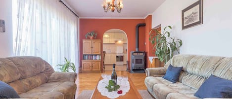 A living room with two sofas, a coffee table with a bottle and glasses, a chandelier, and a kitchen visible through an archway.
