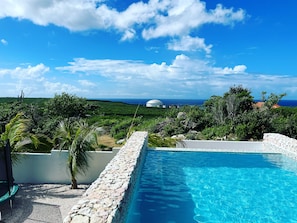 Une piscine bordée d’un mur en pierre, entourée de verdure, avec vue sur l’océan et surmontée d’une structure en forme de dôme.