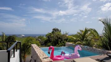 A pool with pink flamingo and unicorn floats, surrounded by palm trees and a stone wall, with a view of the sea and a dome structure in the background.
