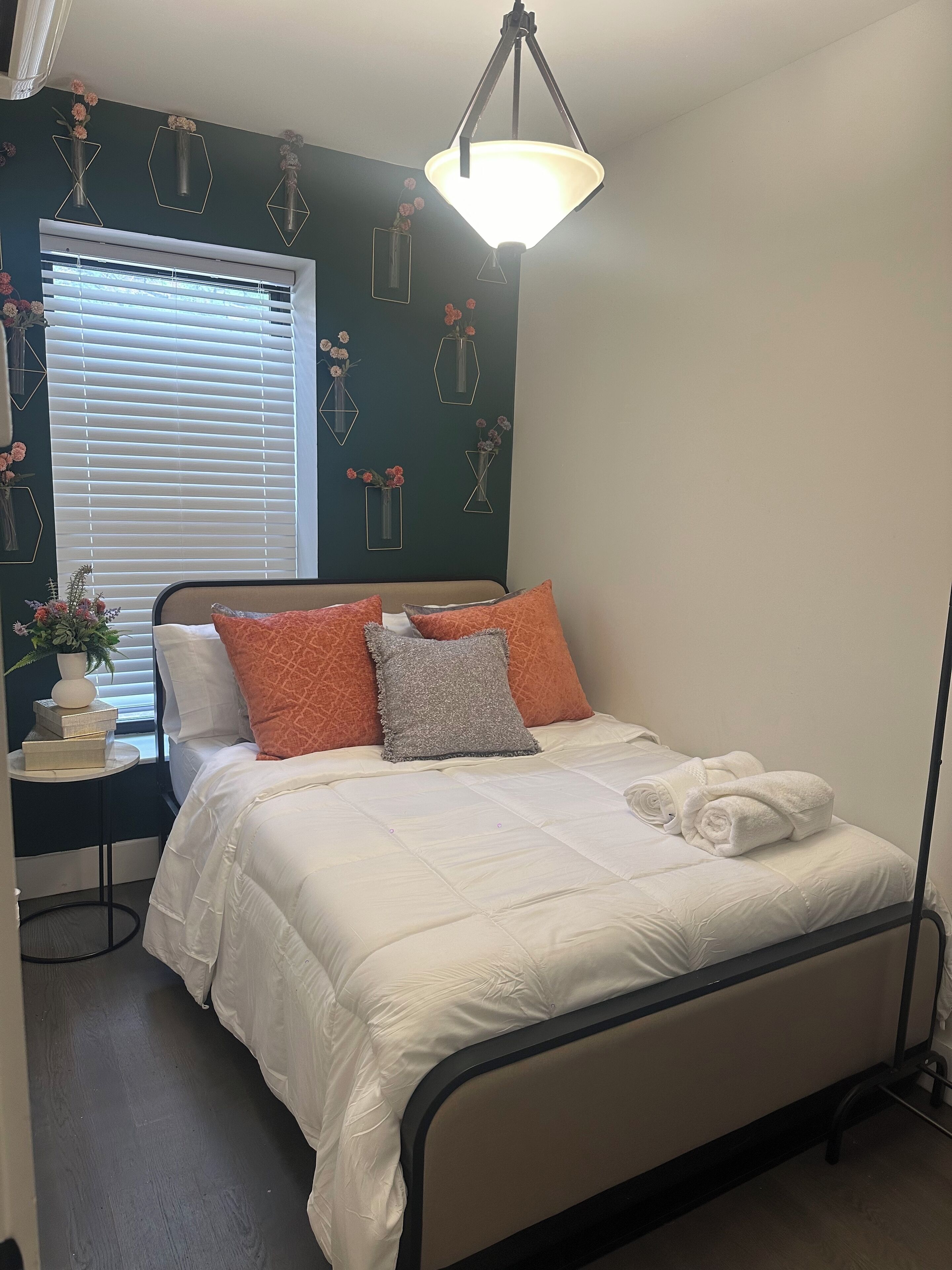 A neatly made bed with white bedding and orange and gray pillows, a bedside table with a vase and books, and a window with blinds.