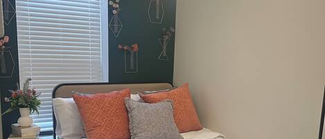 A neatly made bed with white bedding and orange and gray pillows, a bedside table with a vase and books, and a window with blinds.