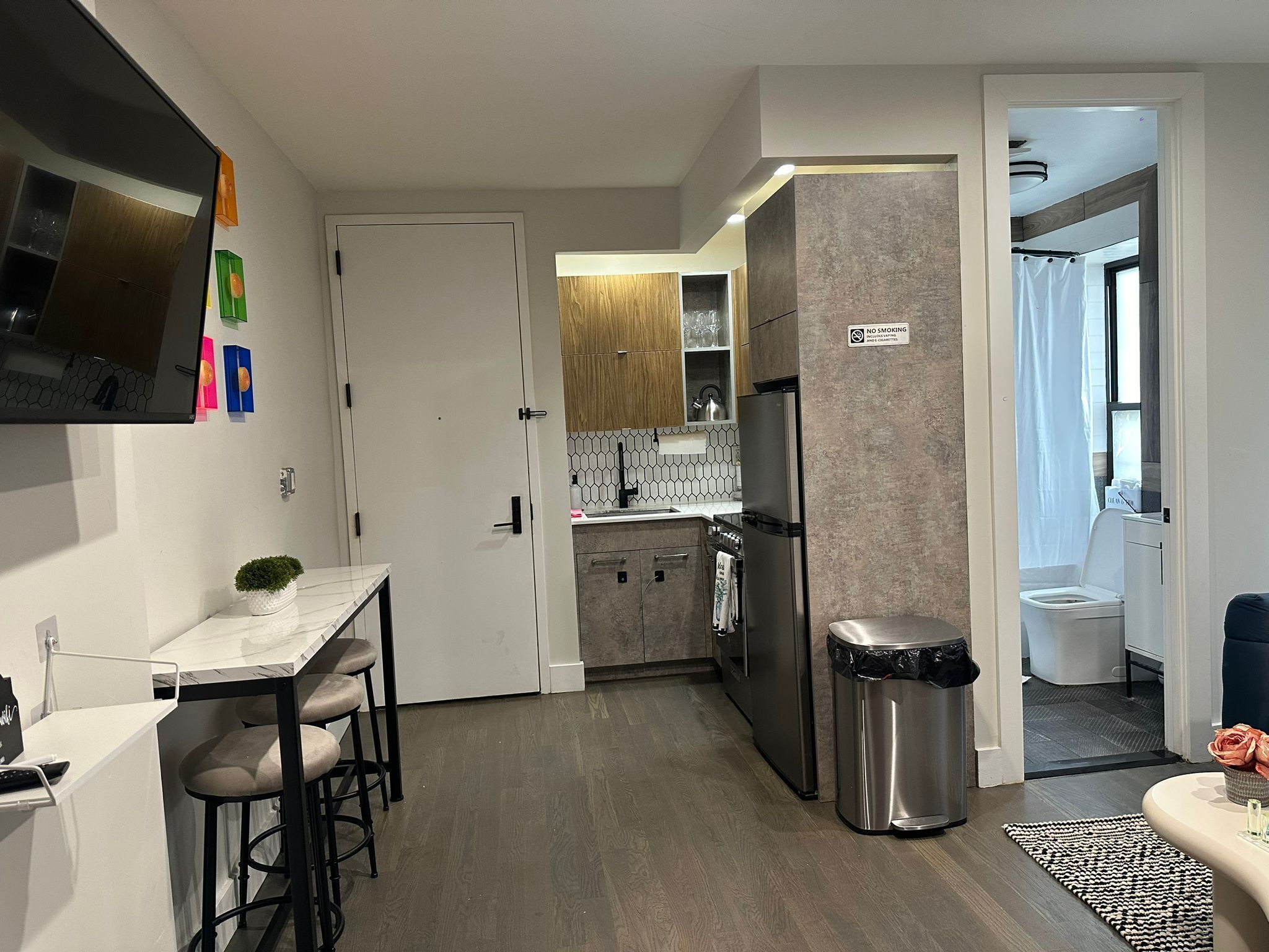 Una cocina moderna con horno empotrado, refrigerador de acero inoxidable y microondas. Hay un pequeño comedor con una mesa blanca y dos taburetes de bar. Un bote de basura está ubicado cerca de la entrada de la cocina.