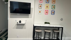 A modern kitchen with a flat-screen TV mounted above a white countertop, a small shelf with kitchen items, and a bar stool area with three stools. - Cozy Stay in NYC!! (New York)