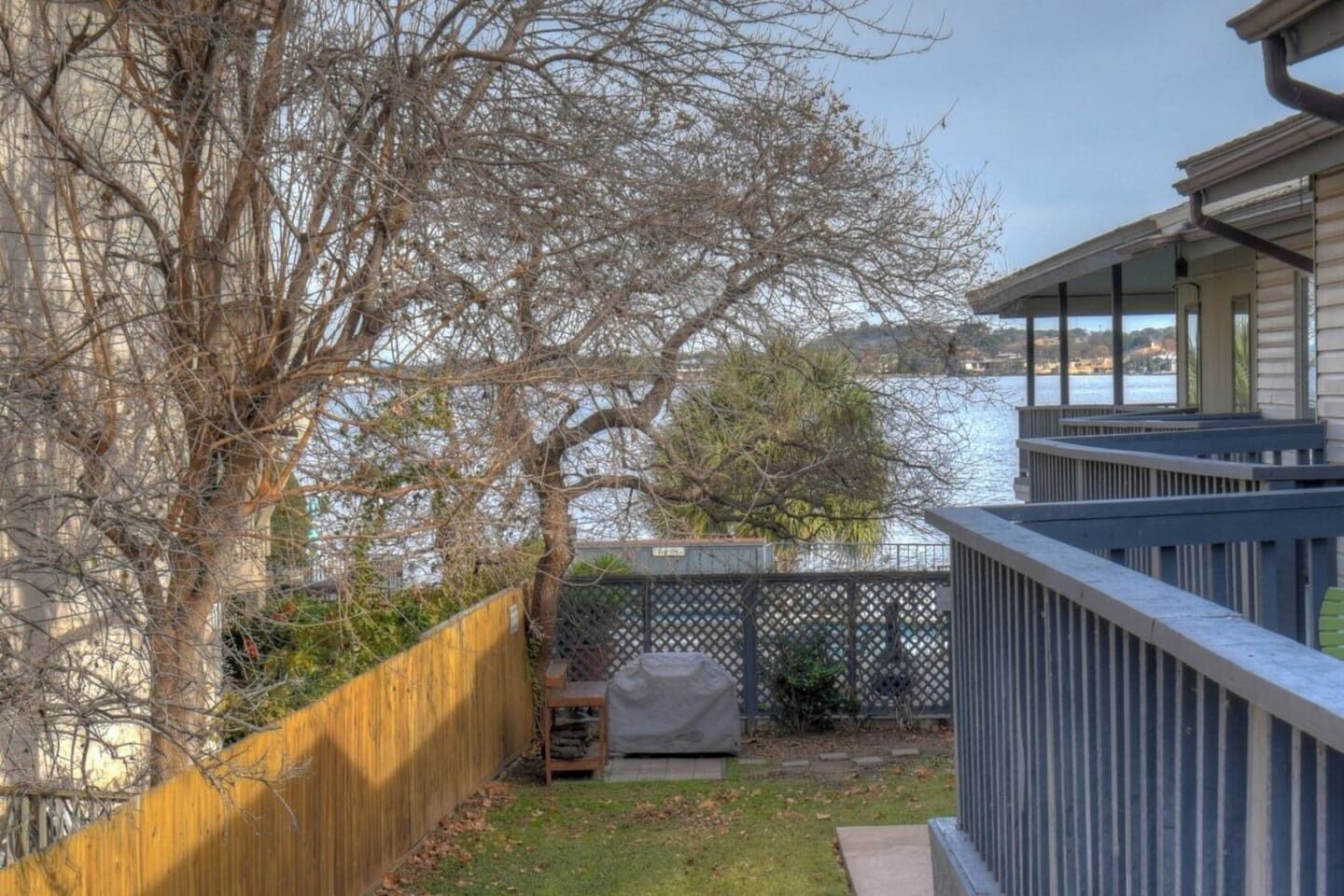 Een achtertuin met een houten omheining, een overdekte barbecue en uitzicht op een waterpartij.