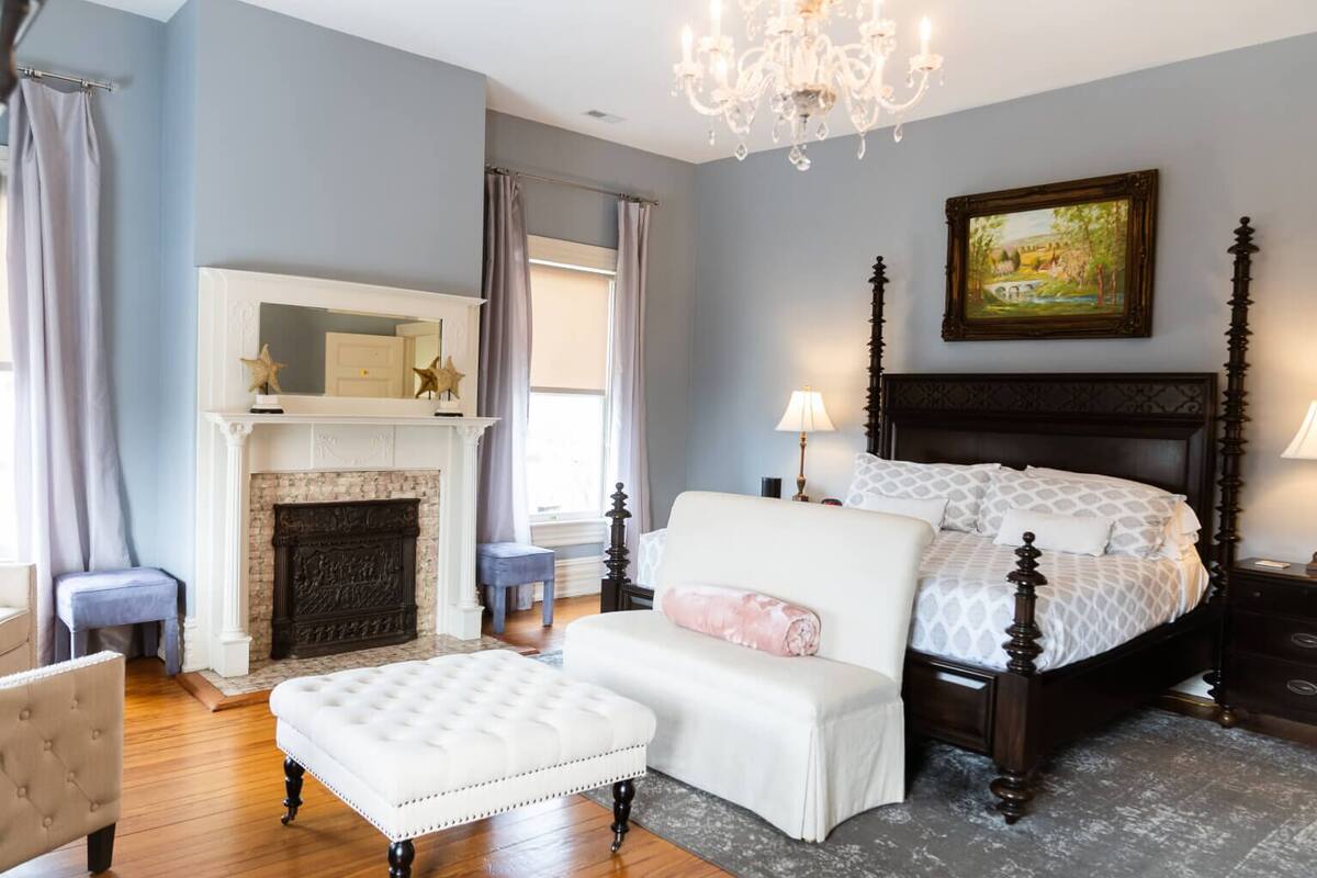 Quarto com lareira, cama com roupa de cama estampada, um banco estofado branco e um candelabro.