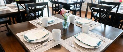Una mesa de comedor dispuesta con platos, cubiertos y tazas blancas, rodeada de sillas.