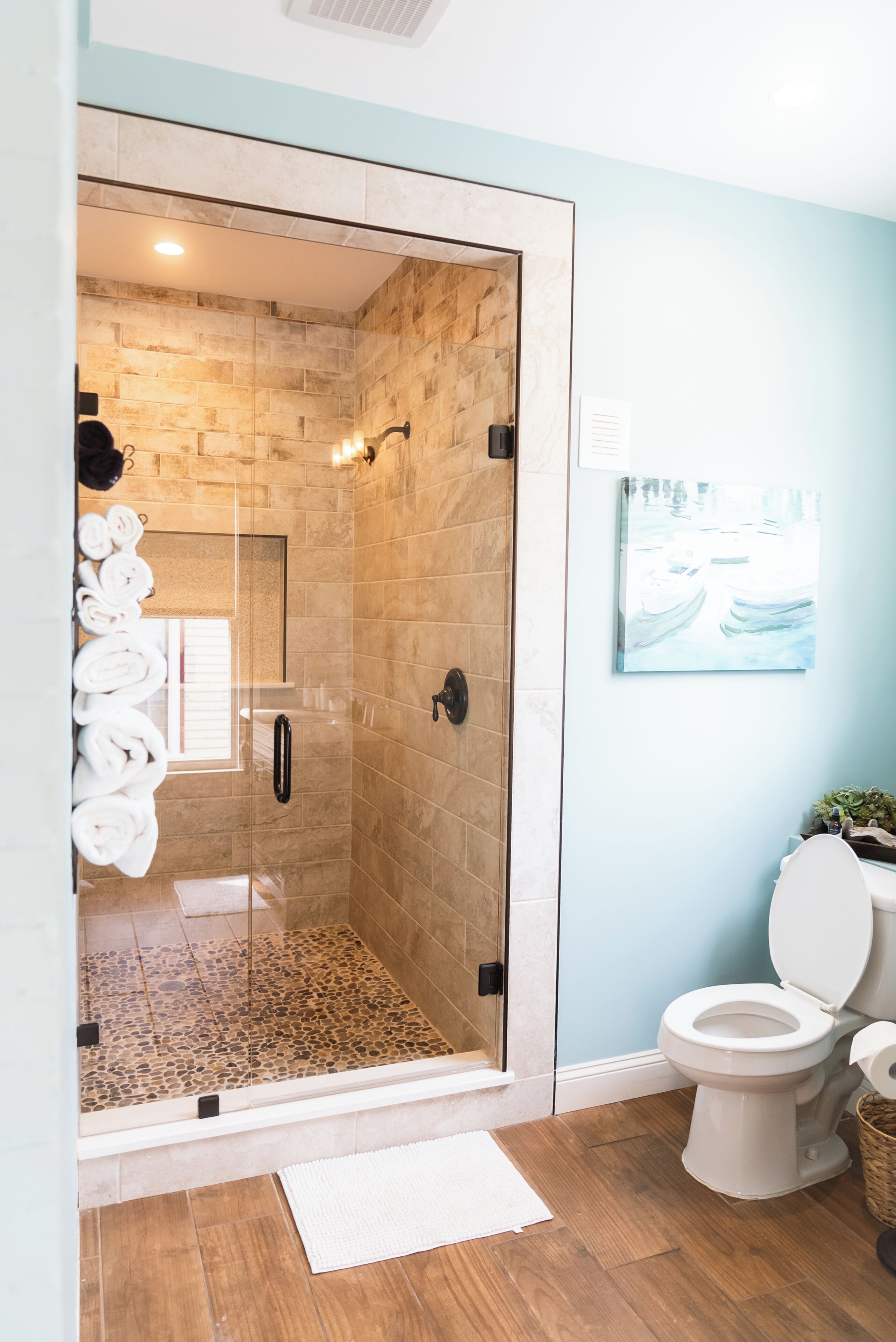 A glass-enclosed shower with stone tiles, a white toilet, and a painting on the wall.