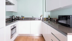 A modern kitchen with white cabinets, a stainless steel range hood, a built-in oven, a microwave, and a large island with a sink. - Native Victoria (London)