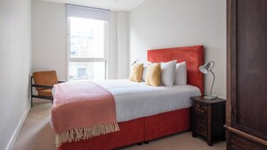 A bedroom with a red bed, white bedding, and a window with a view of buildings. - Native Victoria (London)