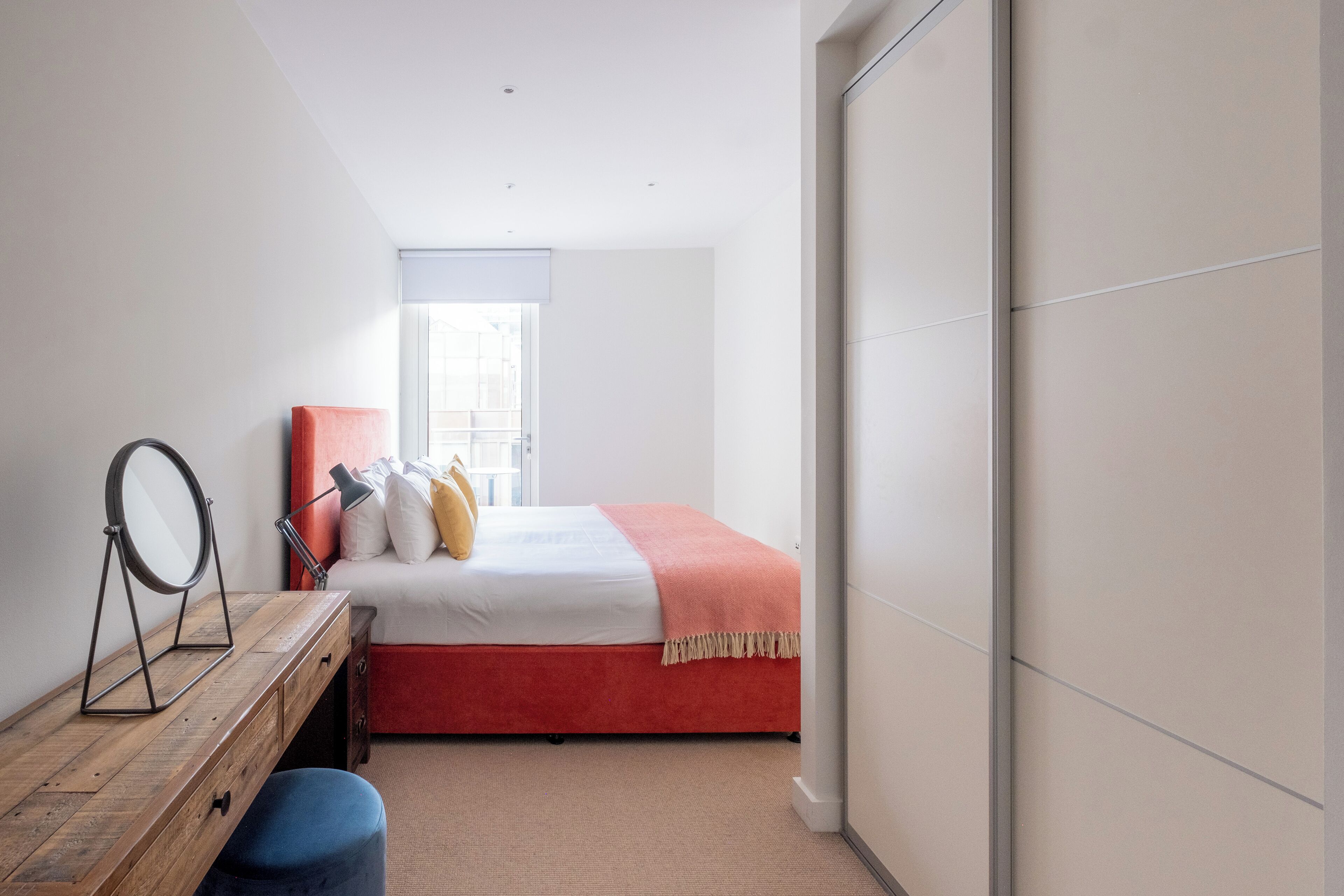 A bedroom with a bed, a vanity with a mirror, a window, and a door.
