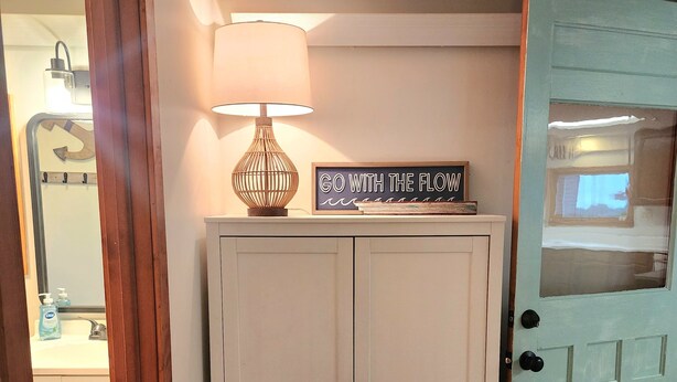 A hallway with a white cabinet, a teal door, a wall-mounted lamp, and a framed sign above the cabinet.