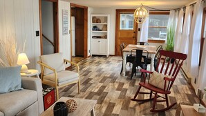 A cozy living room with a wooden floor, a sofa, a rocking chair, a dining table with chairs, and a kitchen area with cabinets and a window. - Charming Lakefront Cottage on Coldwater Lake (Coldwater)