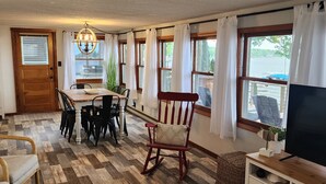 A cozy living room with a wooden floor, a dining table with chairs, a rocking chair, a television, and large windows with curtains. - Charming Lakefront Cottage on Coldwater Lake (Coldwater)