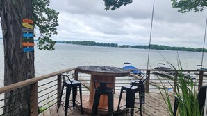 A wooden deck with a round table and chairs, overlooking a lake with boats. A sign on a tree lists amenities like a tiki bar, beach, and fishing.