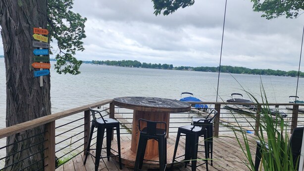 A wooden deck with a round table and chairs, overlooking a lake with boats. A sign on a tree lists amenities like a tiki bar, beach, and fishing. - Charming Lakefront Cottage on Coldwater Lake (Coldwater)