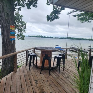 A wooden deck with a round table and chairs, overlooking a lake with boats. A sign on a tree lists amenities like a tiki bar, beach, and fishing. - Charming Lakefront Cottage on Coldwater Lake (Coldwater)
