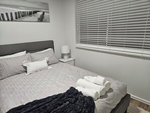 Una cama bien hecha con almohadas y una colcha, una lámpara de noche y una ventana con persianas.
