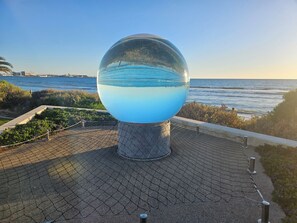 Estructura de vidrio esférica con una base llena de agua, rodeada por un camino pavimentado y follaje, con vista a una masa de agua.