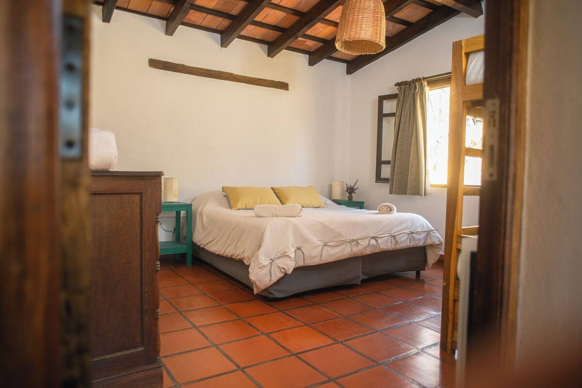 Een slaapkamer met een bed, nachtkastje, een klein tafeltje en een raam met gordijnen.