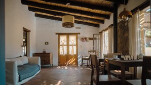 A cozy living room with a sofa, wooden dining table, and chairs. There is a wooden cabinet and a door with glass panels. - Loma Puskana - Hostería (Cachi)