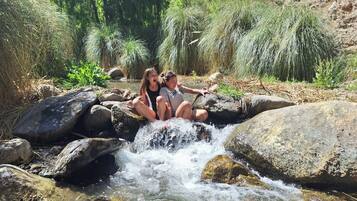 Dos personas sentadas sobre rocas junto a un arroyo rodeado de follaje.