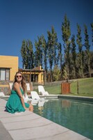 Una persona sentada junto a una piscina con tumbonas y un edificio al fondo.