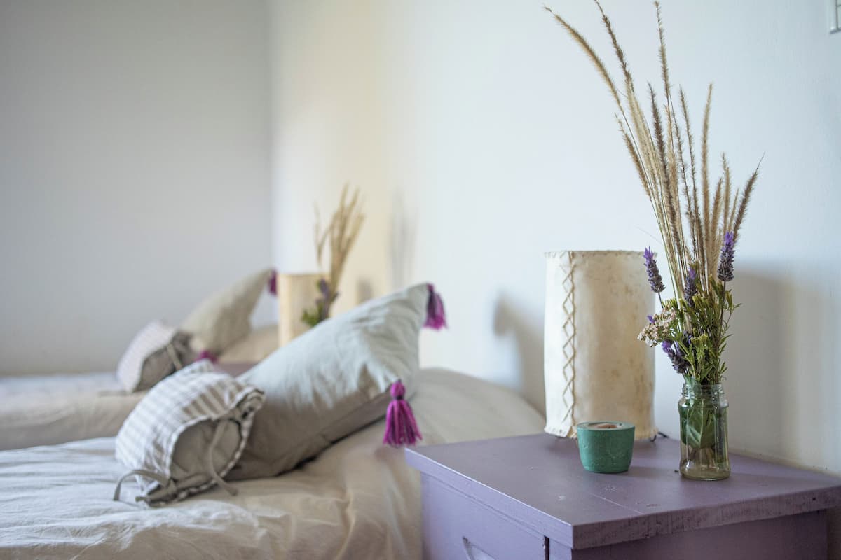 Een slaapkamer met een bed, kussens, een vaas met gedroogde planten en een klein tafeltje met een kaars.