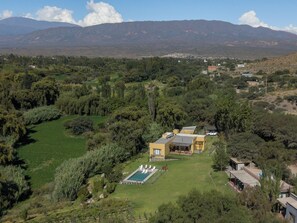 A residential area with a large house featuring a swimming pool, surrounded by lush greenery and mountains in the background. - Loma Puskana - Hostería (Cachi)