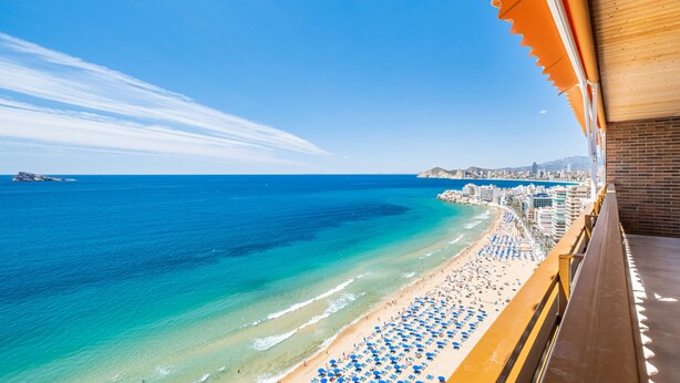 A beach view from a balcony with a wooden railing, overlooking a sandy beach with blue umbrellas and a clear blue sea. - Principado Arena 20 Deluxe Apartment Levante Beach (Benidorm)