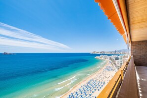 Vista a la playa desde un balcón con baranda de madera, sobre una playa arenosa con sombrillas azules y un mar azul y despejado.