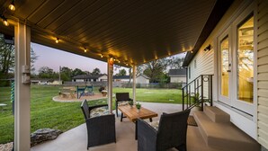 A covered patio with wicker furniture and a wooden table, overlooking a backyard with a lawn and a play area. - Unique Dwtwn+Lake & Trails (Rogers)