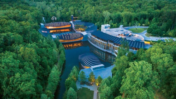 An aerial view of a resort with multiple buildings connected by walkways over water, surrounded by dense forest. - Unique Dwtwn+Lake & Trails (Rogers)