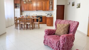 A spacious living area with a dining table and chairs, a kitchen with wooden cabinets, and a patterned armchair.