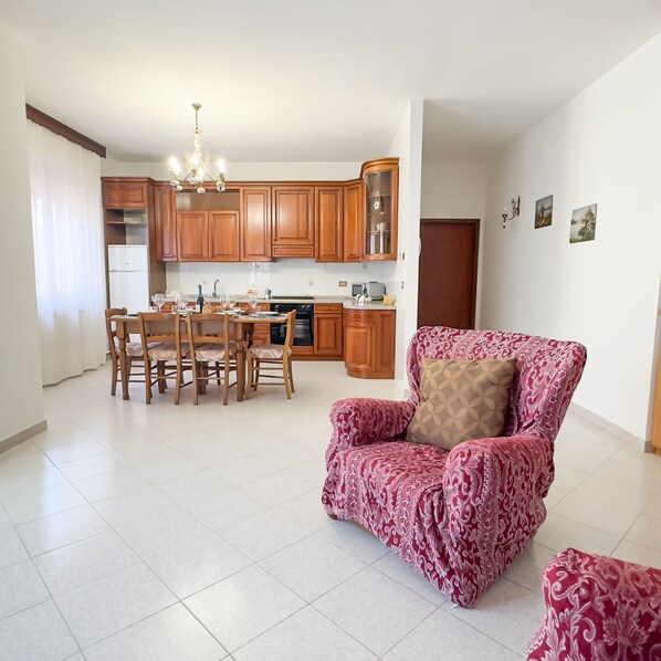 A spacious living area with a dining table and chairs, a kitchen with wooden cabinets, and a patterned armchair.