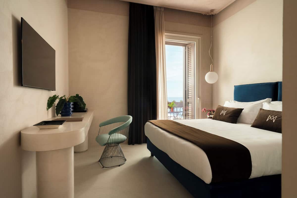 A modern hotel room with a bed, a desk with a chair, a sink, and a large window with a view.