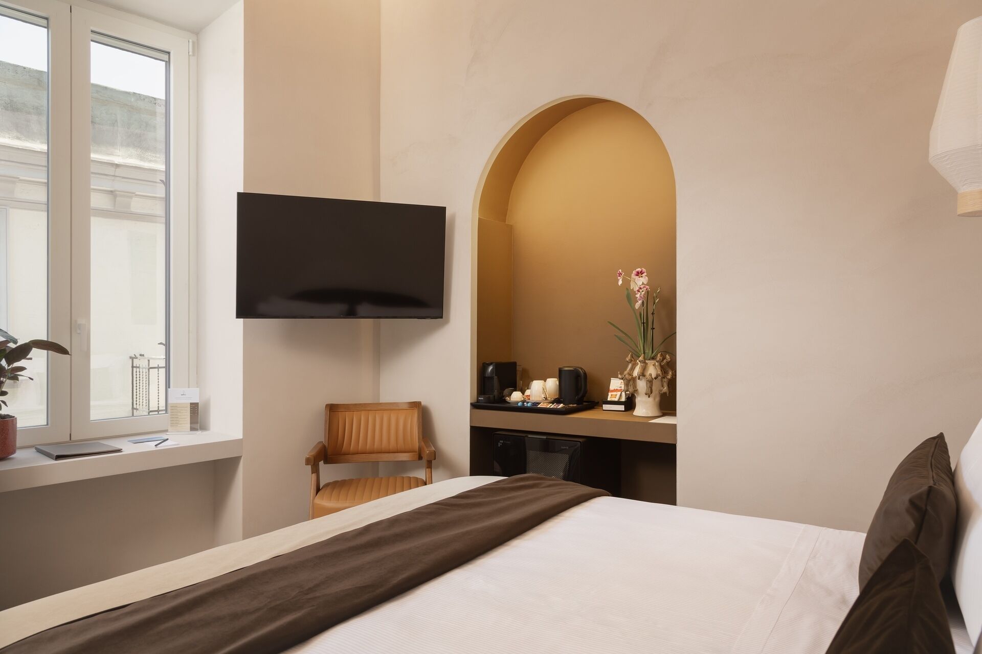 A hotel room with a bed, a desk with a flat-screen TV, a chair, and a potted plant.