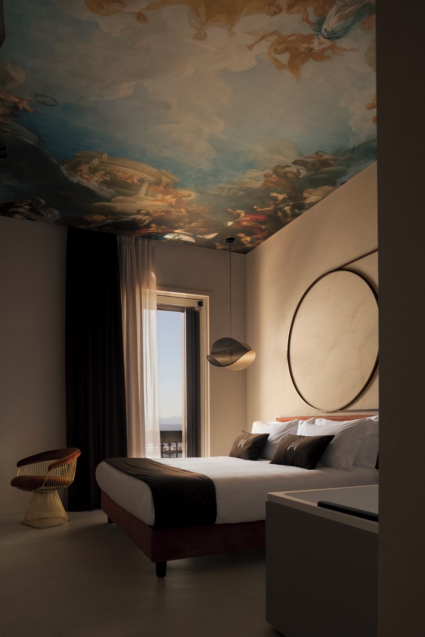 A modern hotel room with a large bed, a round mirror, a ceiling mural, and a view through a window.