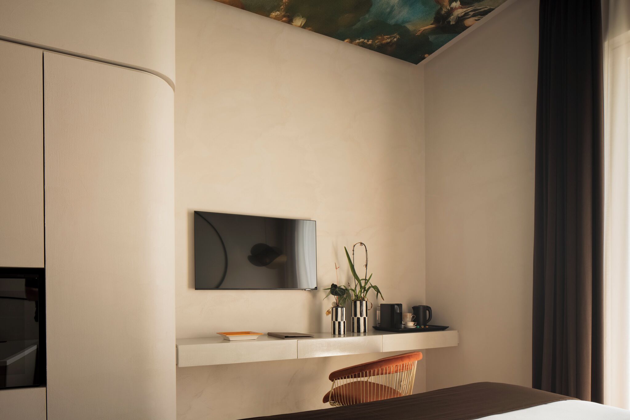 A modern hotel room with a minimalist design, featuring a large flat-screen TV mounted on the wall, a small shelf with decorative items, and a comfortable seating area with a cushioned chair.