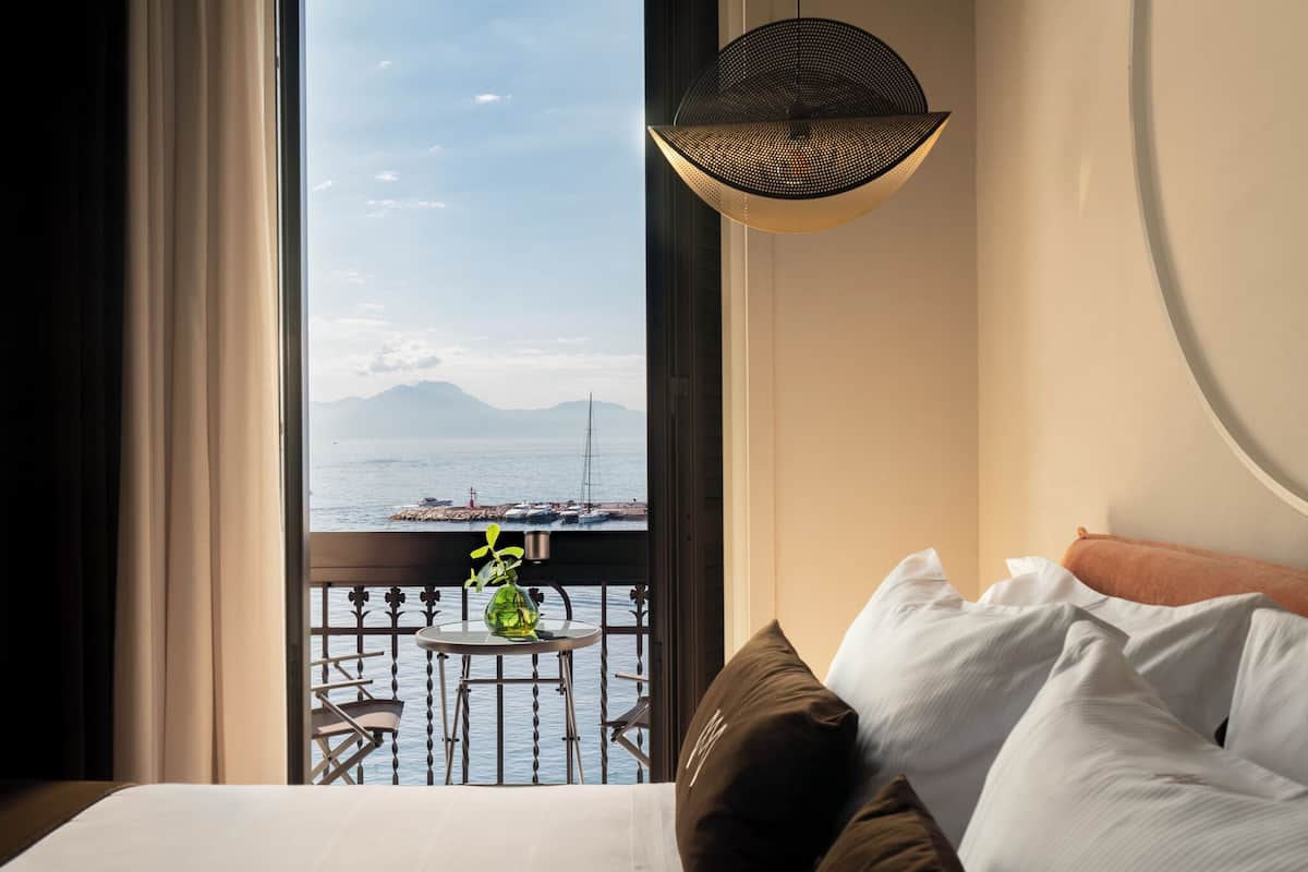 A hotel room with a balcony, a bed with pillows, a hanging light fixture, and a view of the sea and mountains.