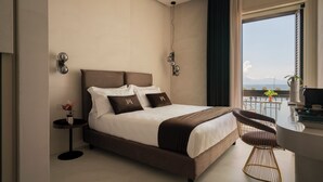 A modern hotel room with a large bed, a small round table, a chair, and a view of the sea. - Demetra Luxury Suites (Naples)