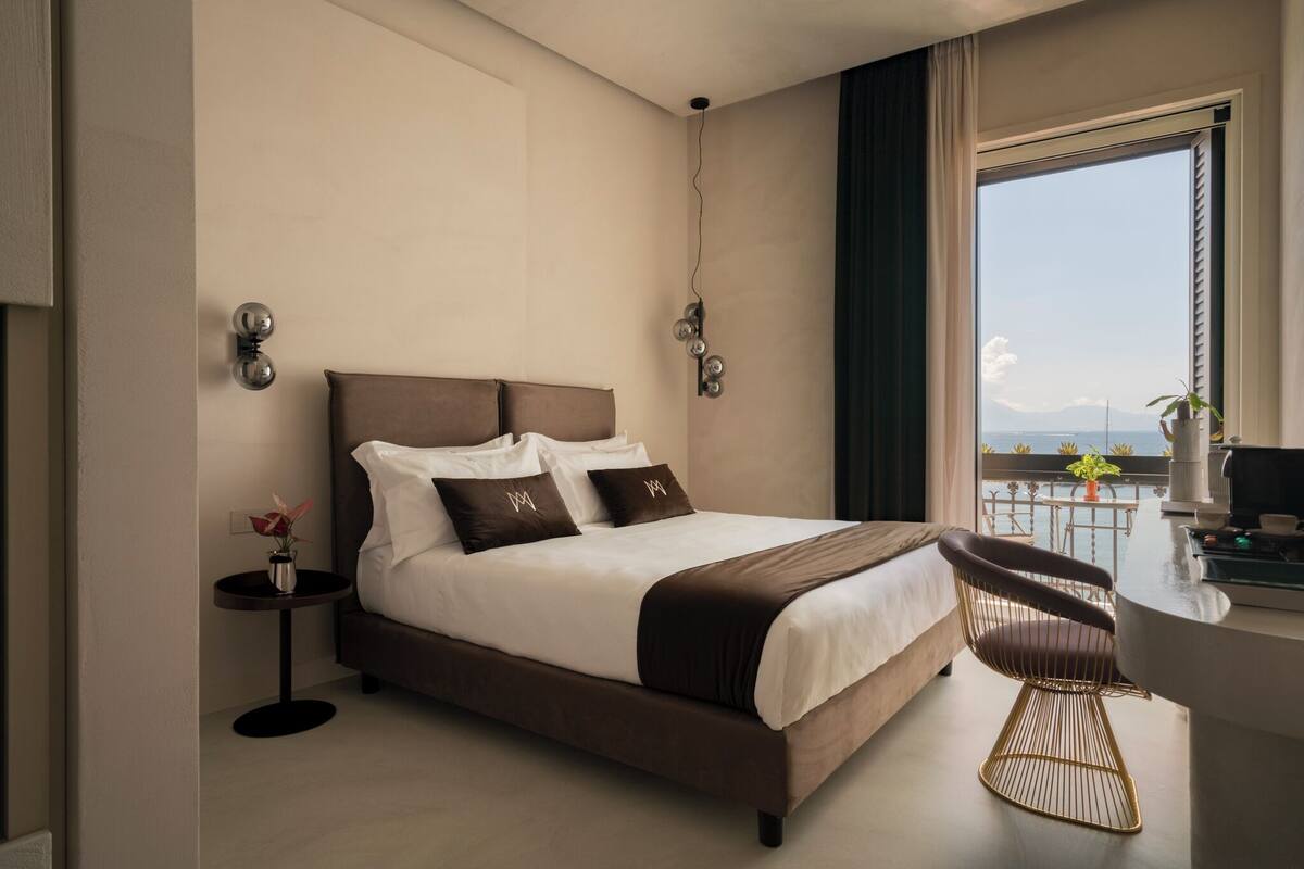 A modern hotel room with a large bed, a small round table, a chair, and a view of the sea.