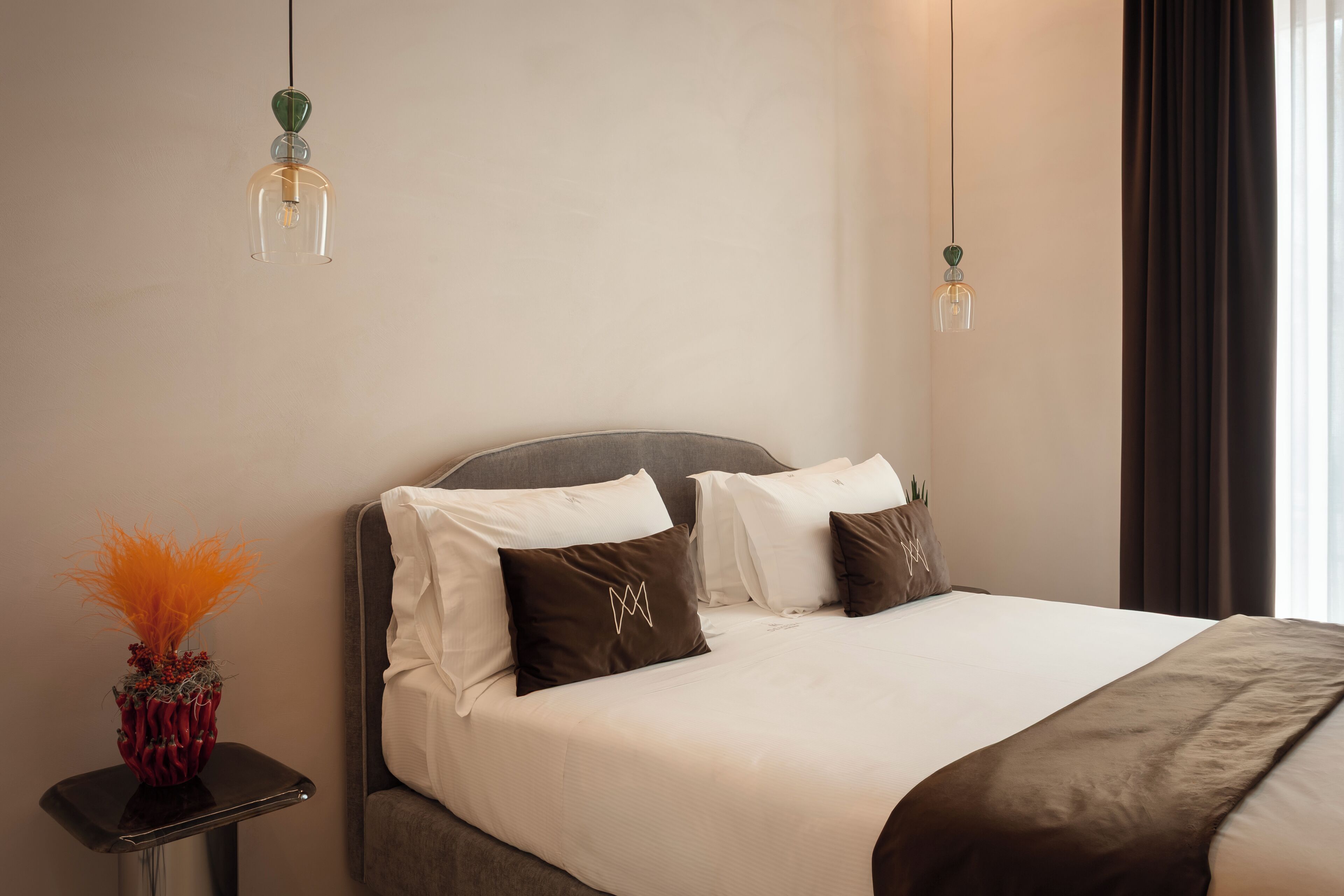 Una cama bien hecha con ropa de cama blanca y marrón, un jarrón con follaje naranja y dos lámparas colgantes.