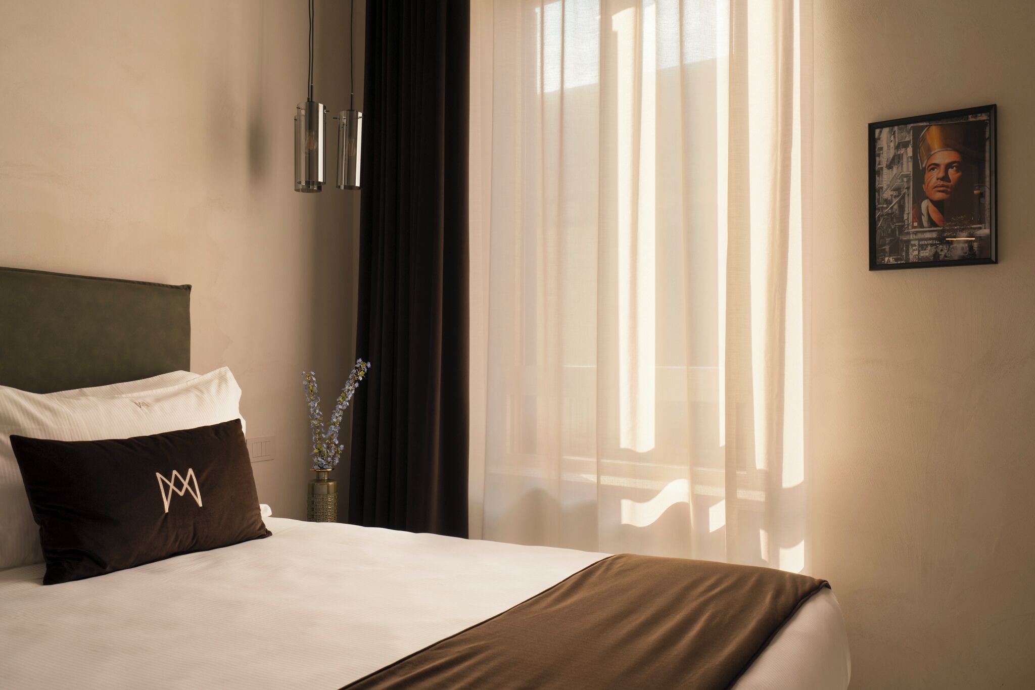 A hotel room with a bed, a headboard, a window with sheer curtains, a wall-mounted light fixture, and a framed picture.