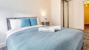 A neatly made bed with a blue blanket and pillows, a bedside table with a lamp, and a door leading to another room. - Immaculate 2-bed Apartment in Birmingham (Birmingham)