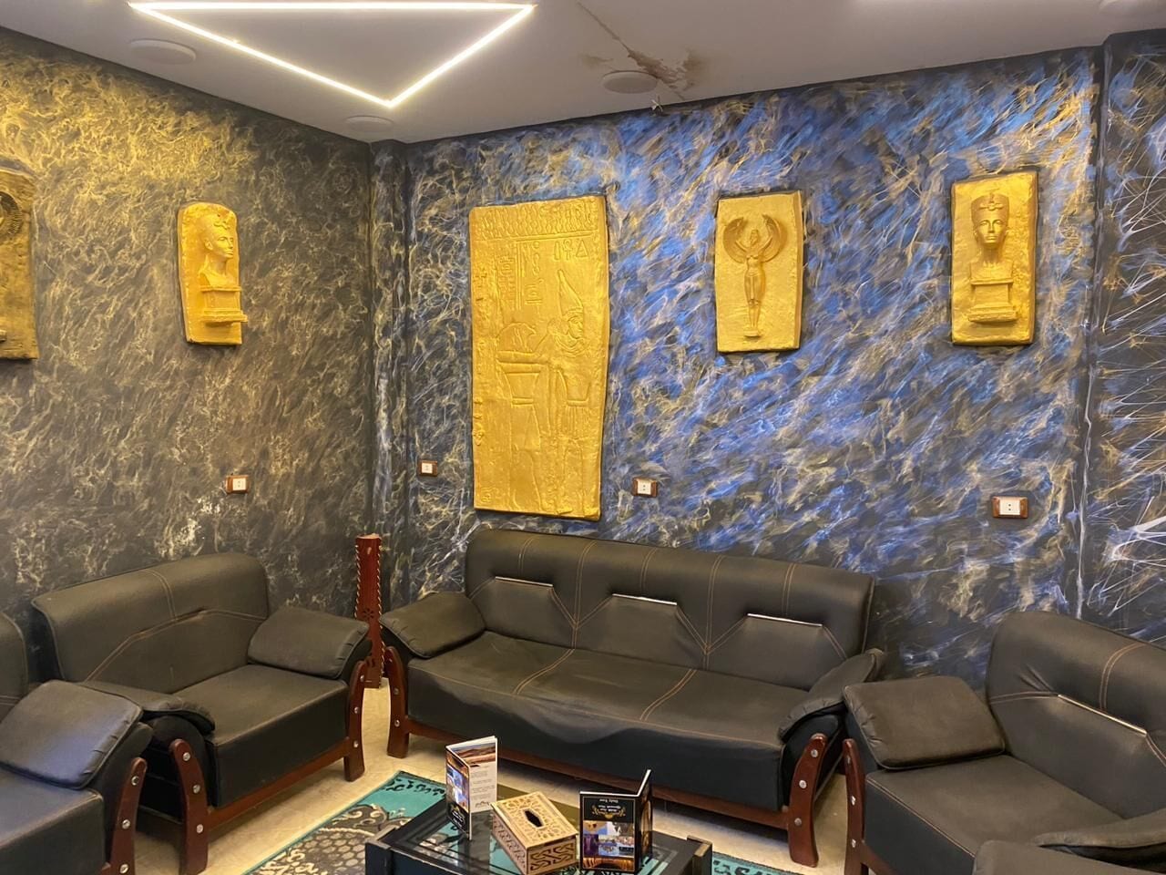Una habitación con pared de mármol azul oscuro, tres sofás de cuero negro y placas doradas.